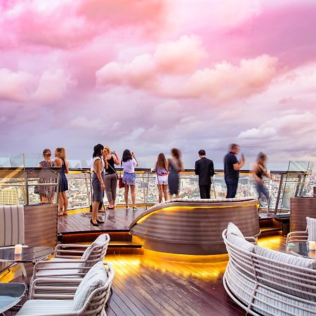Hotel Lebua At State Tower Bangkok Esterno foto The photo depicts a stylish rooftop setting, likely at a bar or lounge. In the foreground, there are elegant, modern seating areas with plush cushions and tables, illuminated by soft lighting. In the background, several people are socializing and enj