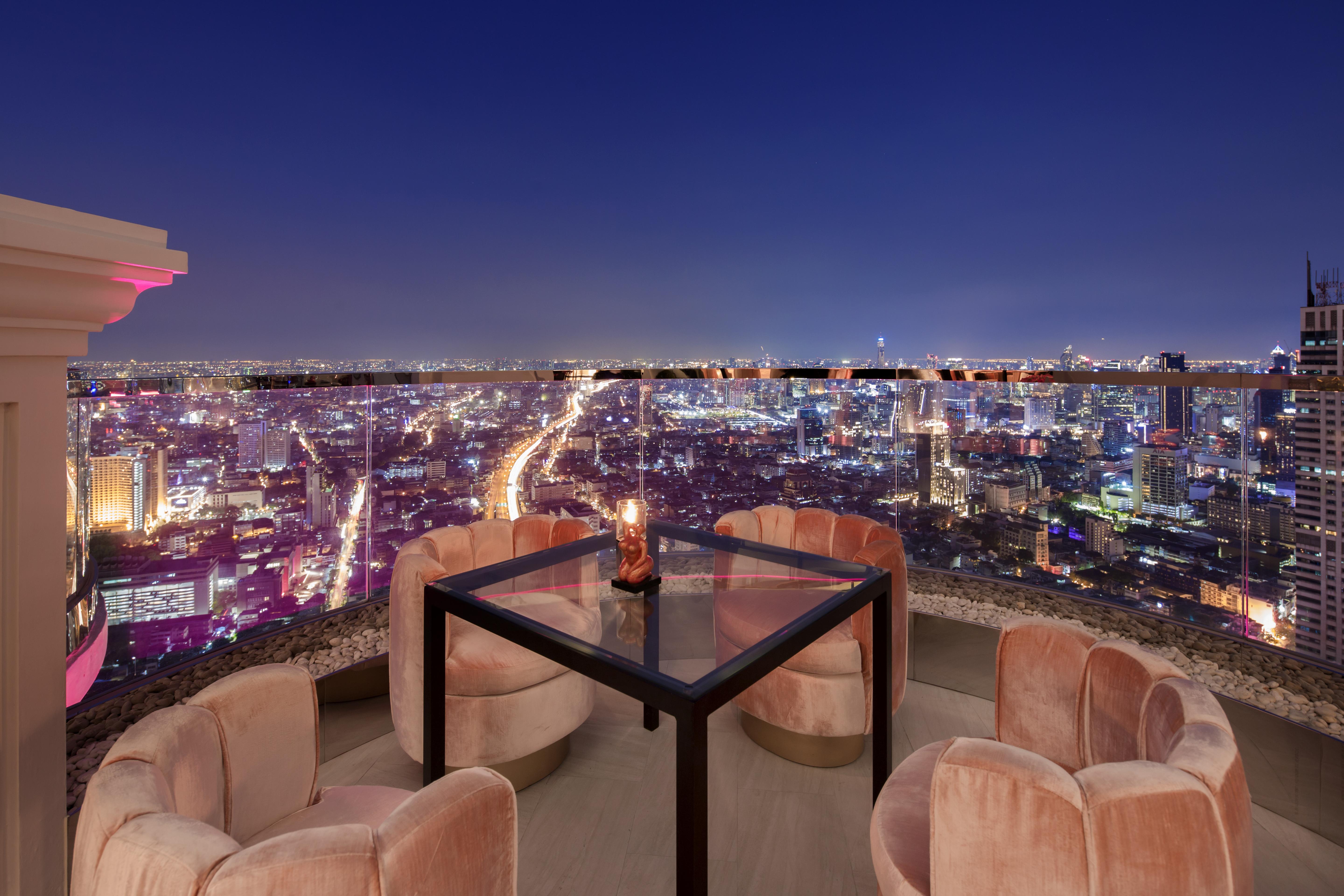 Hotel Lebua At State Tower Bangkok Esterno foto The photo depicts a stylish outdoor seating area on a rooftop, featuring a square glass table surrounded by four plush, circular chairs. The scene is set against a stunning nighttime city skyline, with a multitude of twinkling lights visible in the b