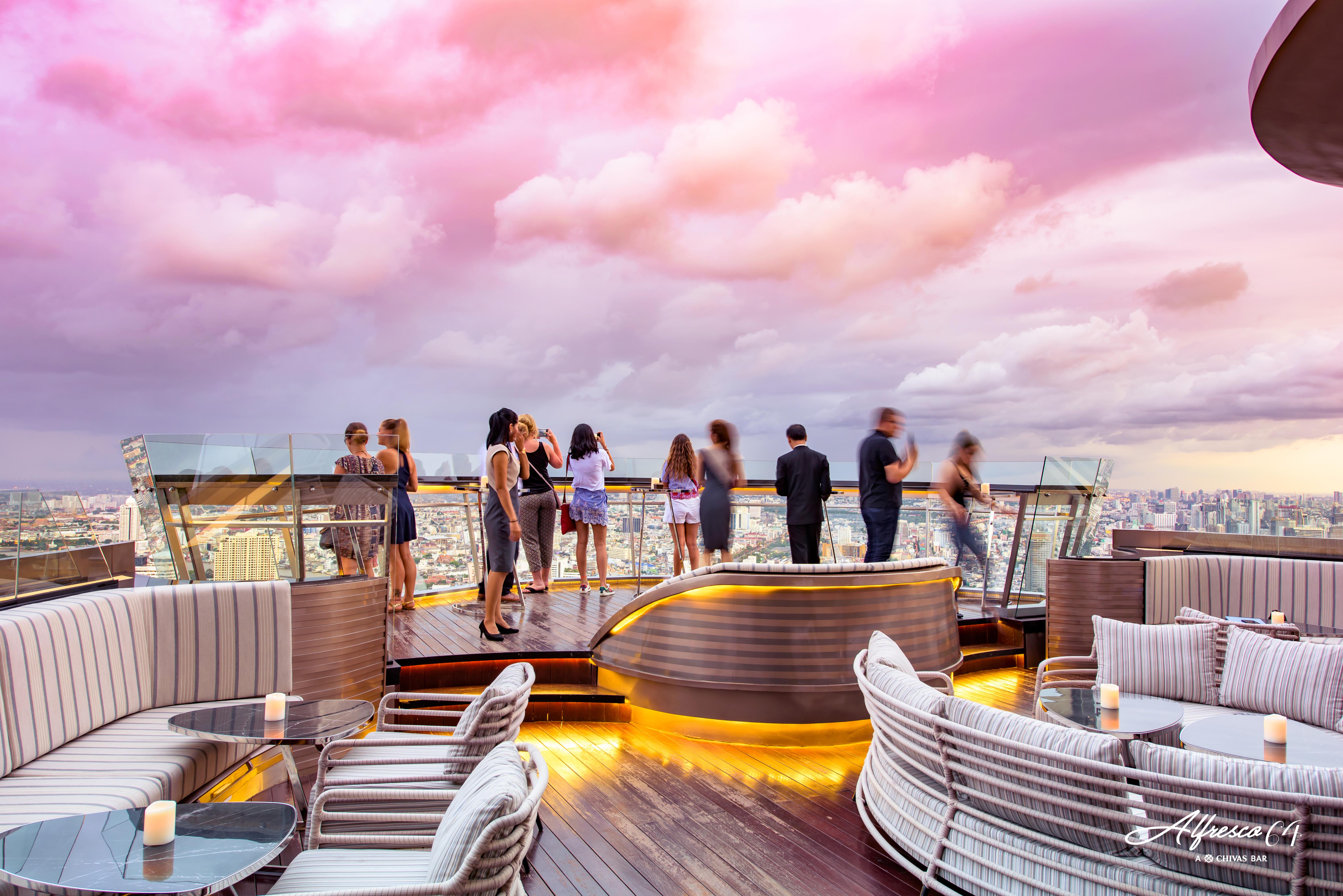 Hotel Lebua At State Tower Bangkok Esterno foto The photo depicts a stylish rooftop setting, likely at a bar or lounge. In the foreground, there are elegant, modern seating areas with plush cushions and tables, illuminated by soft lighting. In the background, several people are socializing and enj