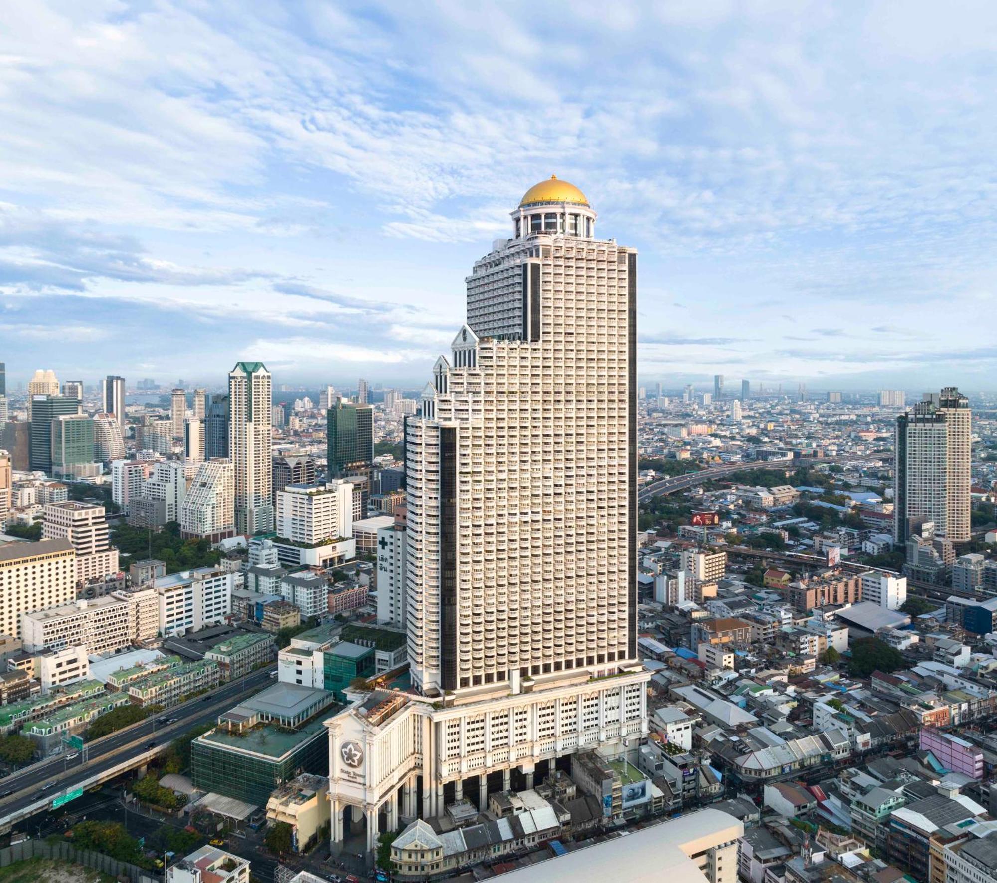 Hotel Lebua At State Tower Bangkok Esterno foto The photo shows a tall, modern skyscraper with a distinctive golden dome on the top. It is situated amidst a bustling urban cityscape, featuring various other buildings of different heights and designs. The surrounding area appears densely populated,